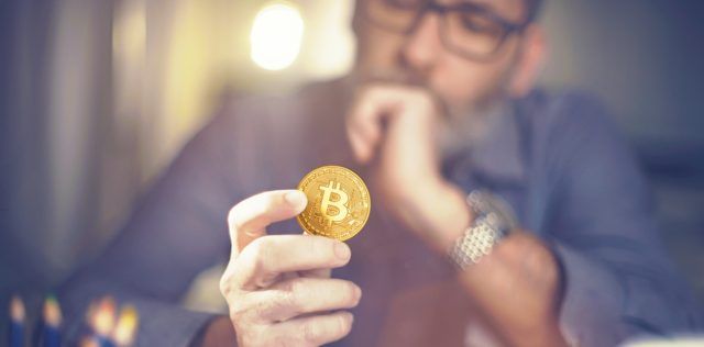 homem segurando uma moeda de Bitcoin com a mão no queixo pensando